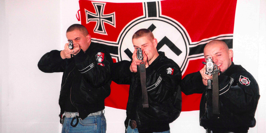 Mitglieder der Hammerskins posieren bewaffnet vor einer Hakenkreuzfahne.